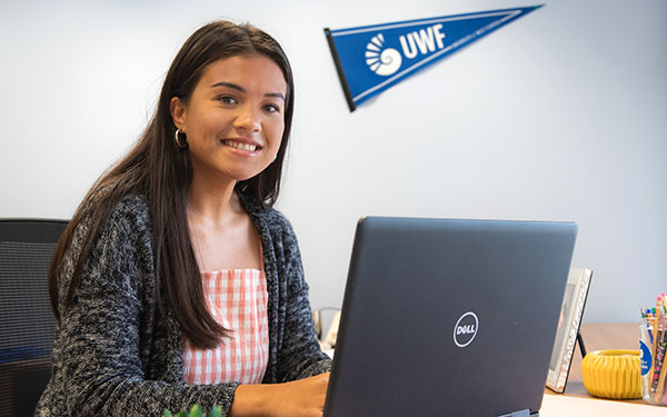 A UWF student smiles while using a laptop as a UWF pennant hangs in the background.