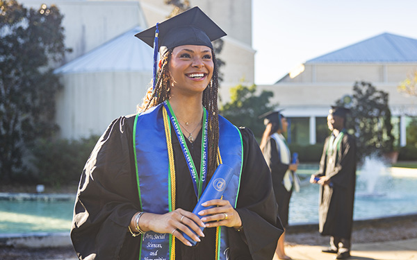 A UWF graduate smiling in cap and gown attire and holding a diploma tube while two people converse in the background while standing in front of a fountain.