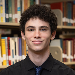Asher Colley indoor headshot in suit and tie attire against a shelf of books in a library.