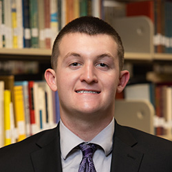Elijah Riffell indoor headshot in suit and tie attire against a shelf of books in a library.