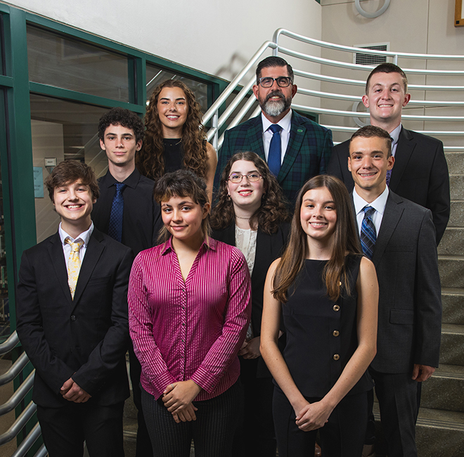 UWF President Manny Diaz Jr. poses with eight students in business attire on a library staircase.