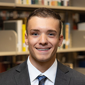 Micaiah Henry indoor headshot in suit and tie attire against a shelf of books in a library.