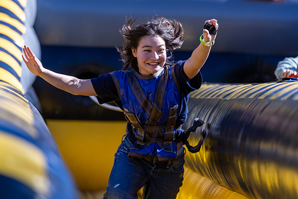 A university student runs joyfully on a colorful outdoor inflatable obstacle course while wearing a safety harness.