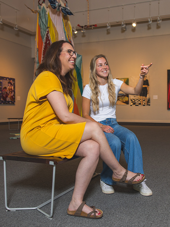 Two students sitting on a bench in an art gallery looking at framed artwork with one student pointing.