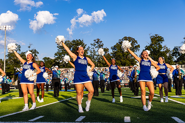 Six cheerleaders in UWF cheer uniforms cheering with pom-poms during a football game halftime performance on a football field with a marching band performing behind them on a sunny day.
