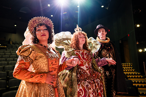 Three theatre students dressed in fancy medieval era attire on an indoor theatre stage.