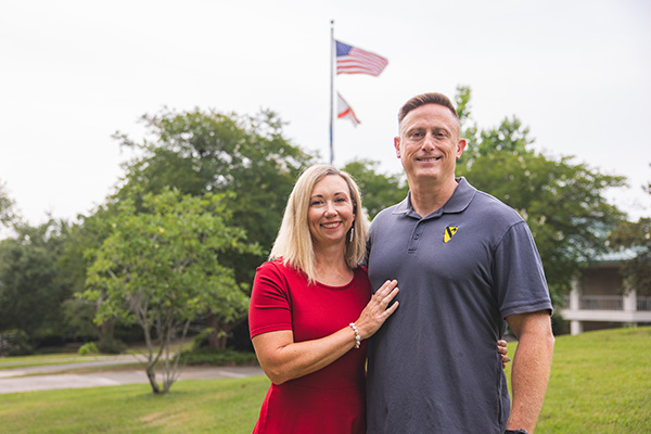 Two people standing outdoors posing close together with trees and a flag pole displaying the U.S. flag and State of Florida flag in the background.