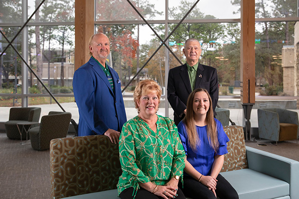 Four members of the Wright family indoors wearing professional attire posing for a group photo with two seated on a couch with two standing behind the couch. A large window is in the background showcasing trees on the University of West Florida campus.