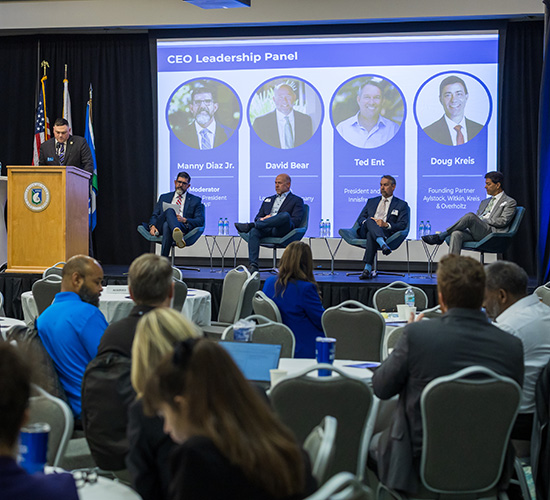 Event speaker in professional attire in front of a podium speaking to a seated crowd with a projected screen displaying CEO Leadership Panel with four speakers seated in front of the display.