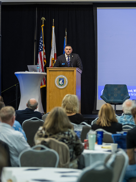 Event speaker in professional attire in front of a podium speaking to a seated crowd with a projected screen displaying Leadership in the Age of AI Conference.