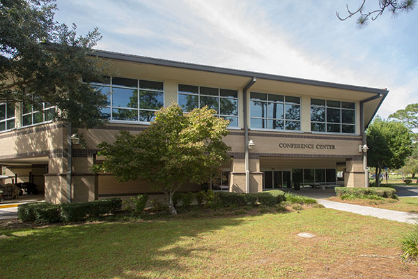 Exterior of the UWF Conference Center, a beige two-story brick building with large upper-level windows, a covered entrance, and landscaped grounds.