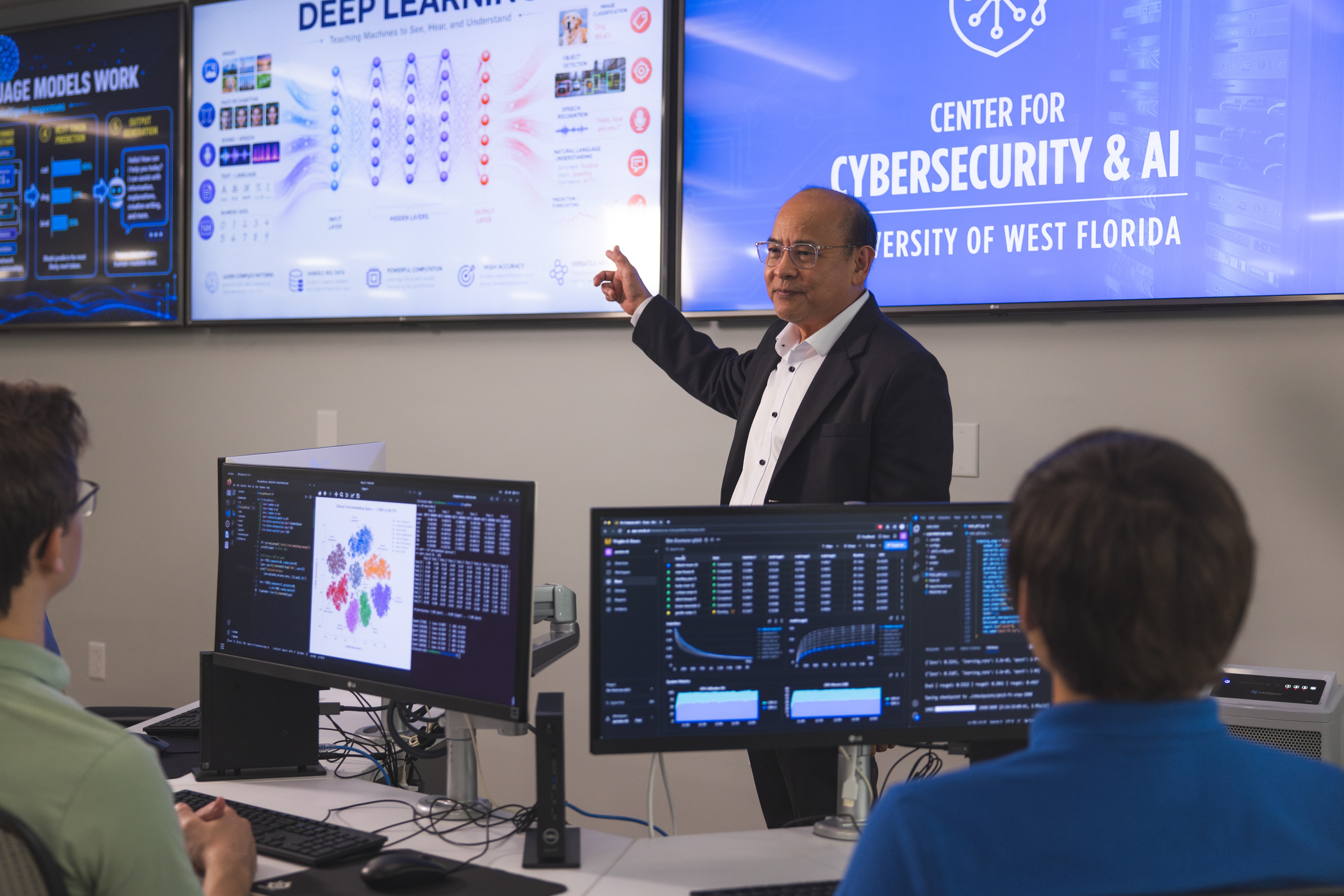 A male instructor stands at the front of cybersecurity lab, pointing upward toward a large wall-mounted display. Two students seated in the foreground with their backs to the camera.