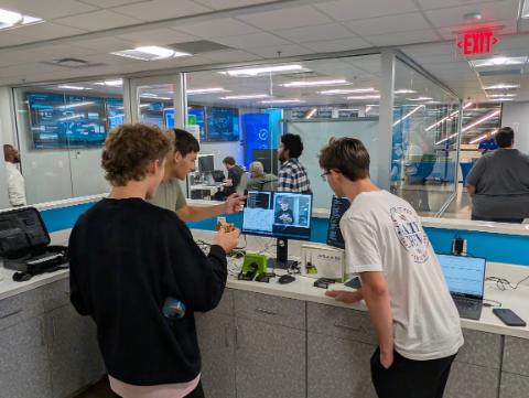 Three young men gather around a computer monitor in a modern glass-walled lab at the UWF Center for Cybersecurity & AI. One student points at a live video feed and code on the screen next to a JetBot AI Kit box and hardware components, while another watches leaning over the counter.