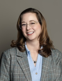 Studio headshot portrait of Dr. Elizabeth Rasnick smiling against a neutral gray backdrop, wearing rimless glasses, a light blue collared shirt, a pendant necklace, and a patterned plaid blazer.