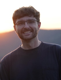A portrait photo of a smiling man with short curly brown hair, a beard, and glasses, wearing a dark short-sleeve shirt. He is photographed outdoors at golden hour, with a soft orange and pink sunset sky and hazy mountain silhouettes in the background.