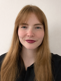 A professional headshot of a young woman (Katarina Kruse) with long strawberry-blonde hair and bangs, fair skin with freckles, and dark red lipstick, wearing a black blouse and smiling gently at the camera against a plain off-white background.