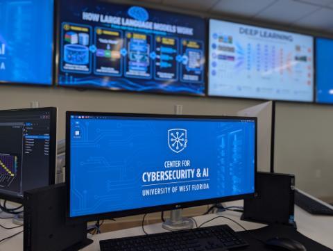 A computer workstation in the UWF Center for Cybersecurity & AI lab features a monitor displaying the center's branded blue wallpaper with the shield-and-circuit logo and the text 