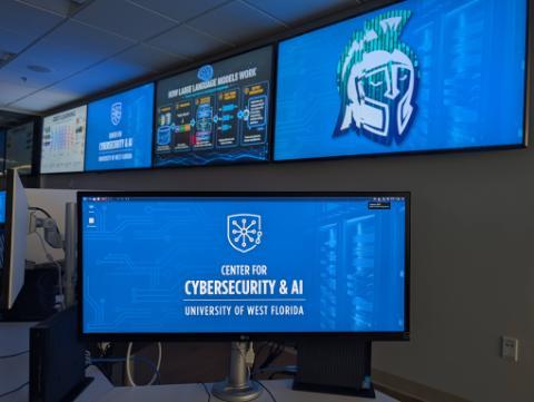 A high-tech workstation in a laboratory environment featuring an LG desktop monitor in the foreground displaying the Center for Cybersecurity & AI - University of West Florida wallpaper. In the background, a large wall-mounted video wall displays slides on Deep Learning, an infograph titled 