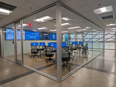 Interior view of the UWF Center for Cybersecurity and AI lab, seen through floor-to-ceiling glass walls. The room features rows of desktop workstations with monitors displaying the Center for Cybersecurity and AI logo, modern ergonomic chairs, and a large video wall showing Center branding and a 