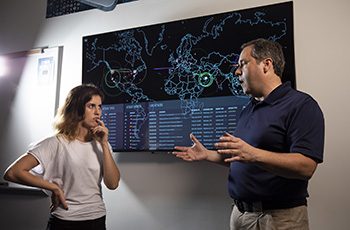 Instructor and student standing in front of a wall monitor displaying a world map of cybersecurity information.