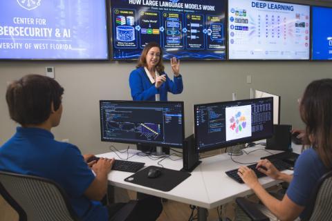 Dr. Elizabeth Rasnick gestures while presenting in a tech lab at the UWF Center for Cybersecurity & AI. Two students sit at workstations with code and data visualizations displayed on their monitors, while digital wall screens behind her illustrate 'How Large Language Models Work' and 'Deep Learning.'