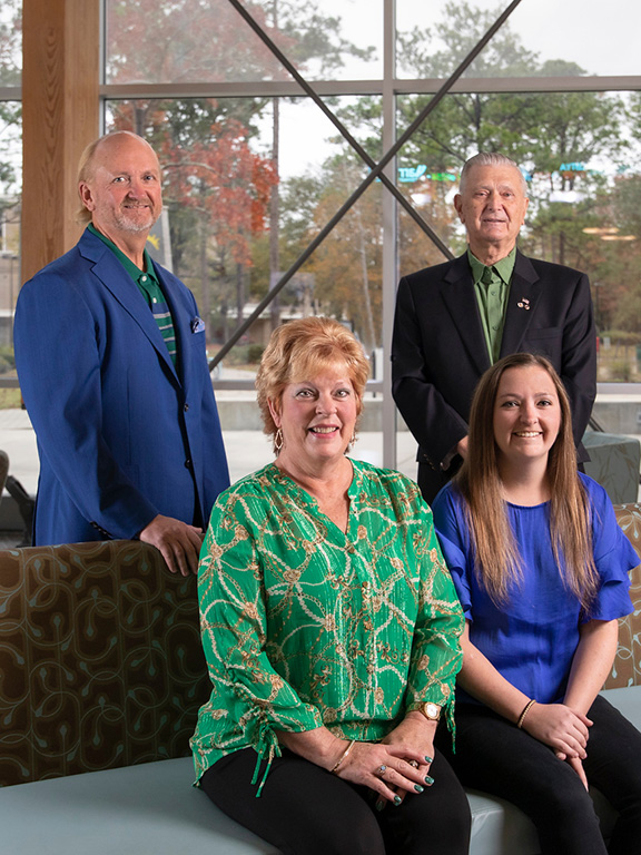 Four members of the Wright family indoors wearing professional attire posing for a group photo with two seated on a couch with two standing behind the couch. A large window is in the background showcasing trees on the University of West Florida campus.