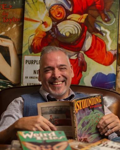 A professor sitting at a desk surrounded by colorful art and magazines.