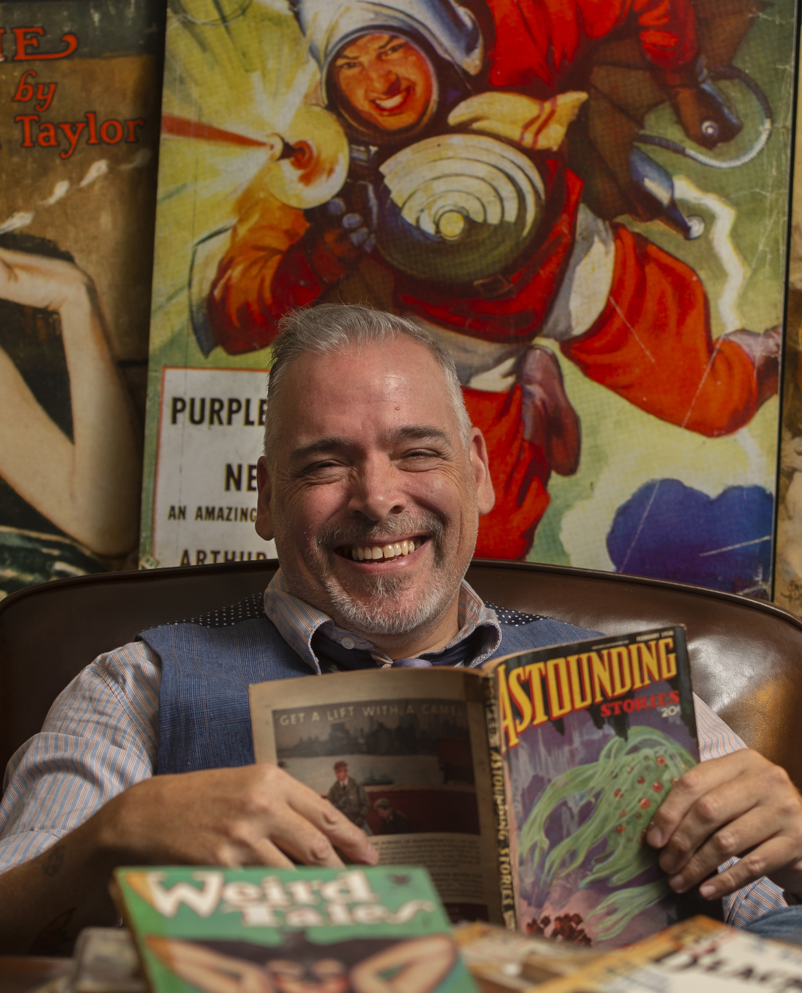 A professor sitting at a desk surrounded by colorful art and magazines.