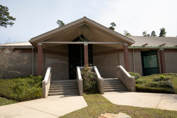 Exterior front view of the Margaret Jane Smith Archaeology Institute building at the University of West Florida. The building is a modern, single-story structure with a neutral brick facade and a wide, gabled roofline. Three sets of concrete stairs with handrails lead up from a paved walkway to a recessed main entrance. A distinctive circular, wheel-shaped metal ornament is mounted at the peak