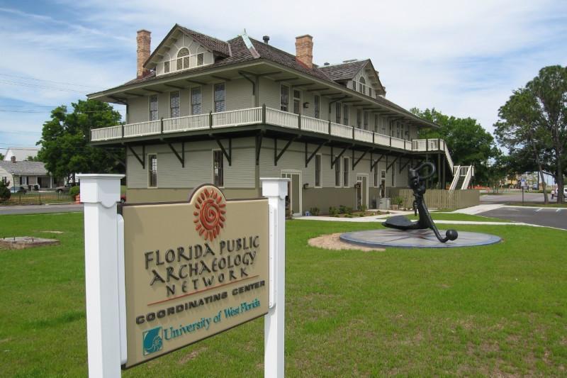 Two-story historic building with wraparound veranda on the second story. A large ship anchor and a Florida Public Archaeology Network Coordinating Center sign are displayed in front of the building.