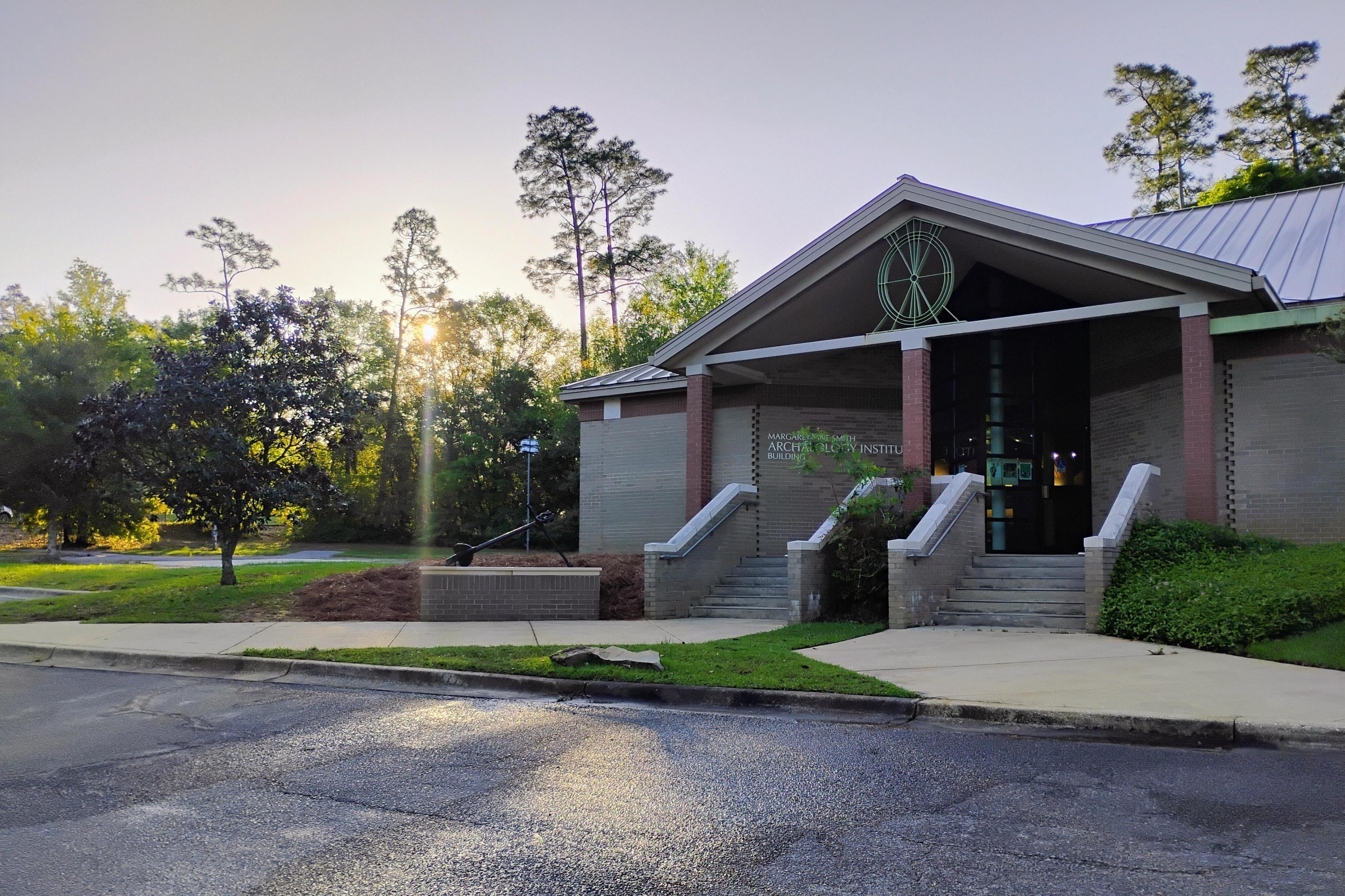 The front of the Margaret Jane Smith Archaeology Institute Building. Two sidewalks and sets of stairs lead to a entry with a peaked roof featuring a turtle medallion the sun is rising in the background