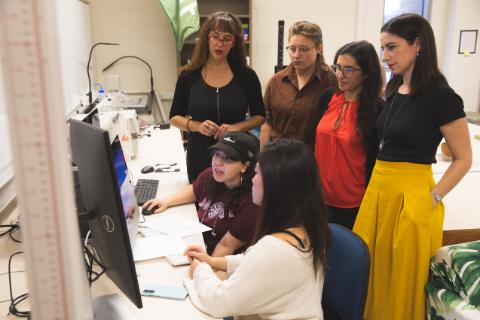 A group of individuals are gathered consulting with two more individuals working on a computer