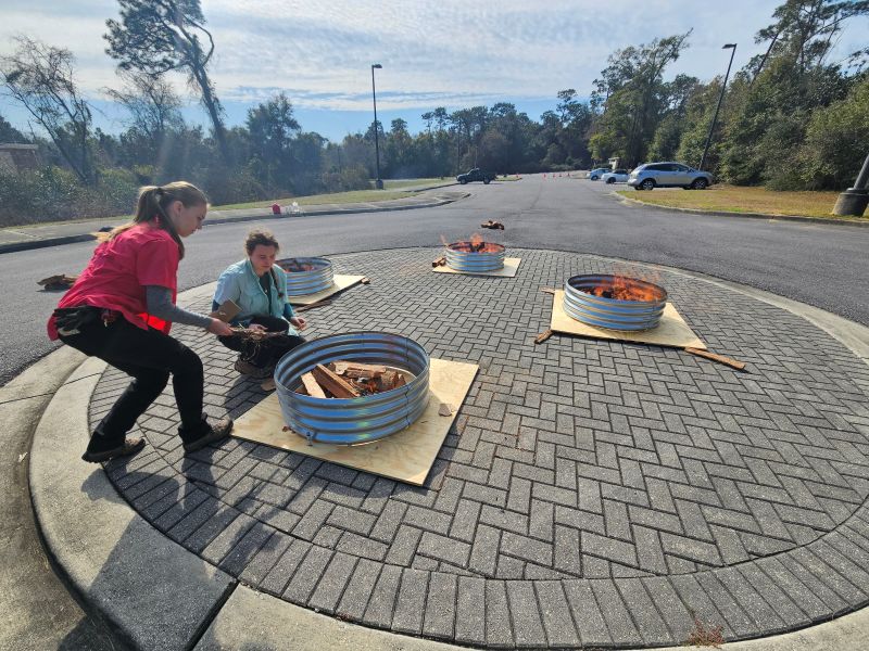 Two women tend to four corrugated metal fire rings placed on plywood sheets in a paved cul-de-sac. Several of the rings contain active fires with orange flames, while the woman in the foreground in a pink shirt adds kindling to a newly lit ring. Other parked vehicles and trees are visible in the background under a bright, clear sky