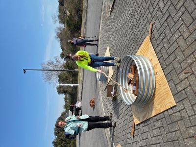 A group of four people prepare wood in corrugated metal fire rings on sheets of plywood in a paved parking lot under a clear blue sky. A person in a neon yellow shirt uses a long stick to arrange wood inside one of the rings.