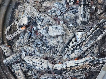 A top-down, close-up view of smoldering logs and glowing embers inside a corrugated metal fire ring. The wood is mostly charred black and covered in grey ash, with small orange flames and glowing red heat visible in the crevices