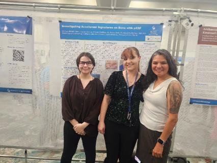 Three women stand smiling in front of a research poster titled 