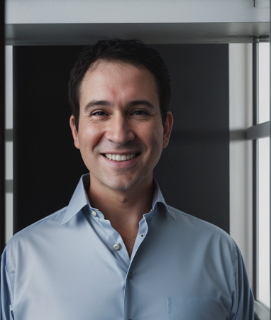 A smiling dark haired man wearing a button front collared shirt