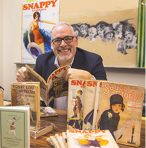 Man at a desk holding a magazine and surrounded by magazines on the desk