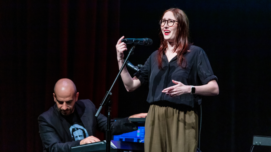 Two musicians on stage, one is sitting and the other is standing while singing