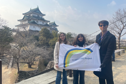 Three people stand in front of a Japanese shrine holding a banner.