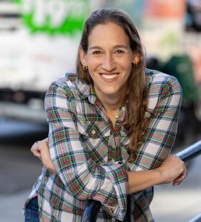 A headshot of a visiting speaker with long light brown hair in an outdoor setting wearing a plaid buttoned shirt.