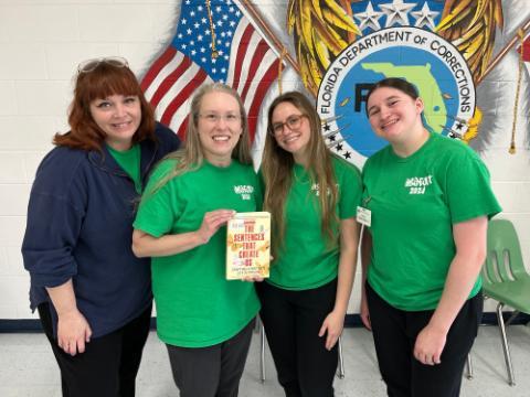 A faculty member and three students in green t-shirts holding a book.