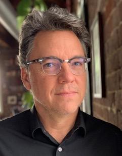 A man with glasses and a black shirt stands in front of a brick wall. - Image credit to Madeleine Casad.