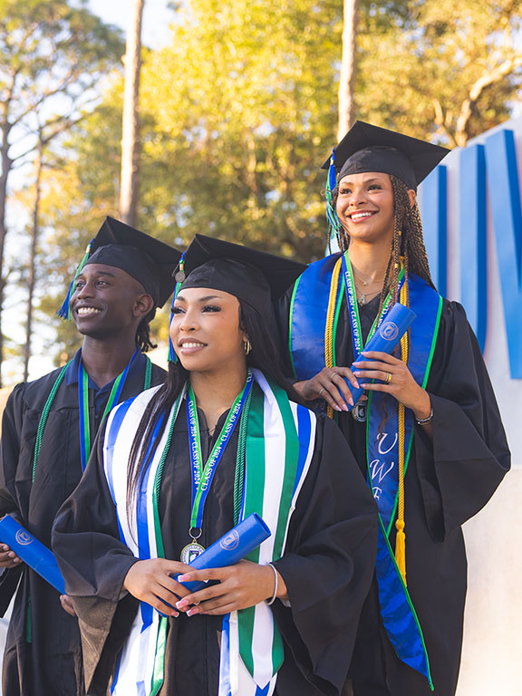 three UWF graduates posing in front of the UWF sign at the campus entrance wearing their graduation regalia