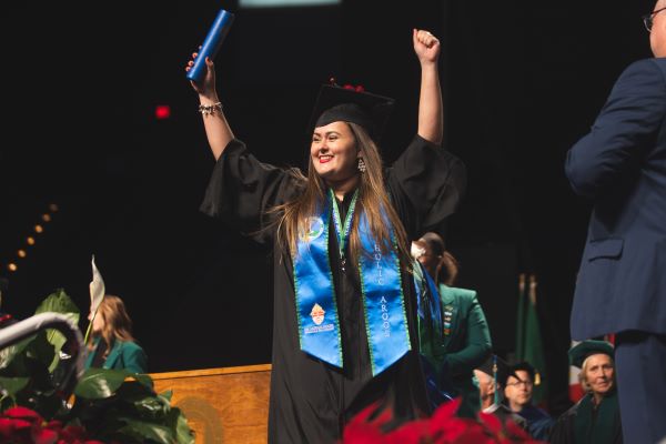 Student in graduation regalia walking across the stage with arms held up in a victorious pose