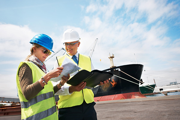 Two shipyard workers wearing hard hats and reflective vests reviewing documents at a port with a large cargo ship in the background.