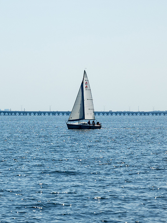 A sailboat carrying three people crosses the water with a long bridge in the background.