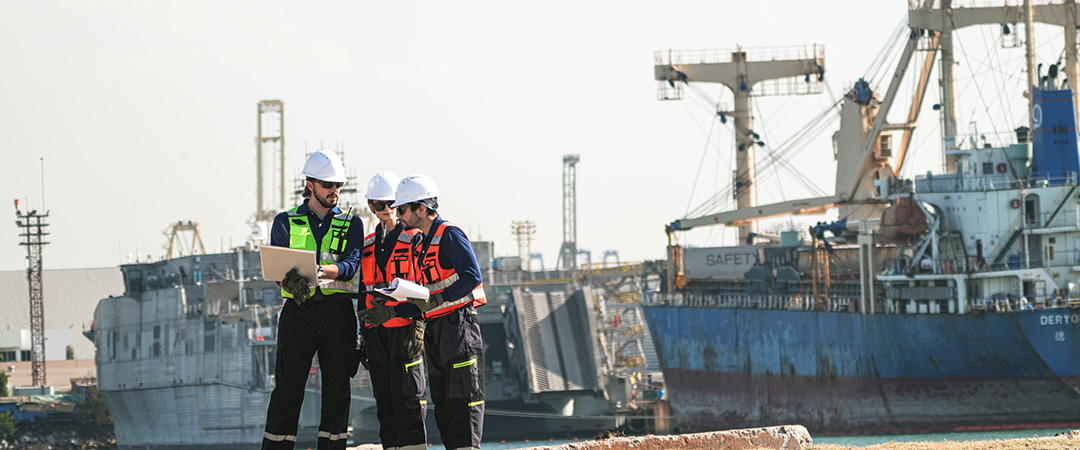 Three professional port workers stand on the gravel ground of a busy industrial port fully equipped with safety gears including hard hats and high-visibility vests while collaborating using a laptop and clipboard.