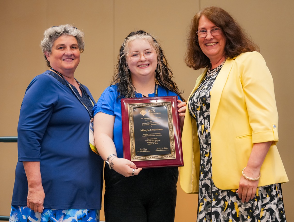 Three women stand together; the one in the middle holds a plaque.