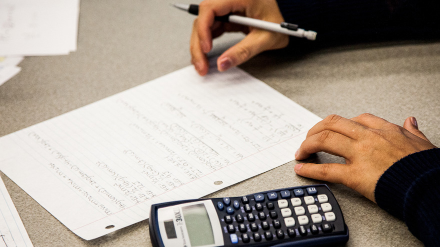A right hand holding a pen next to a piece of paper with written notes and a calculator.